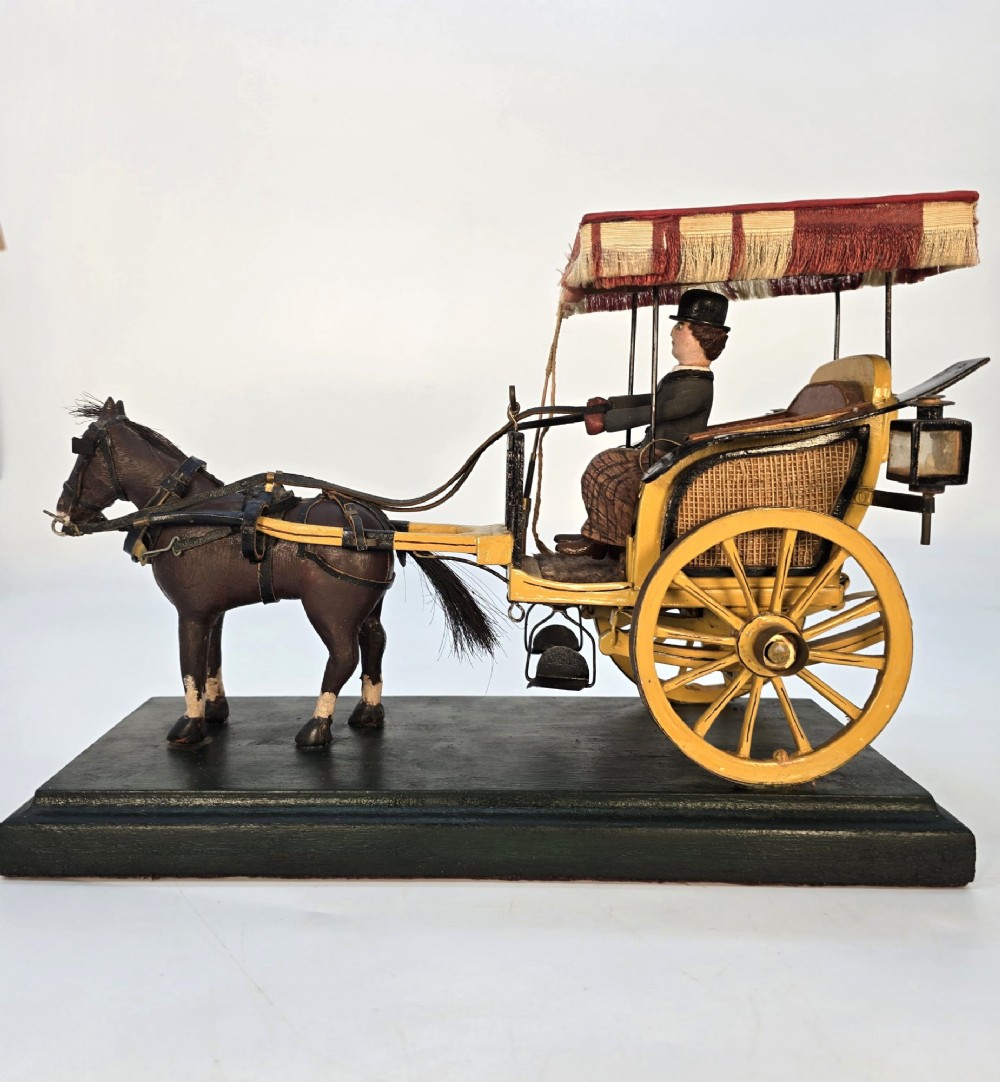 A hand-crafted 1928 wooden folk art model featuring a brown horse attached to a yellow two-wheeled chaise carriage with a driver figure under a striped canopy, mounted on a dark wooden base. View IV