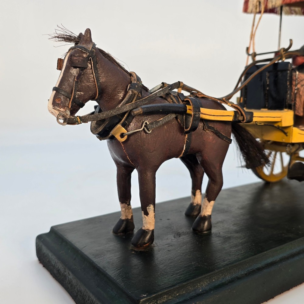 A hand-crafted 1928 wooden folk art model featuring a brown horse attached to a yellow two-wheeled chaise carriage with a driver figure under a striped canopy, mounted on a dark wooden base. View V