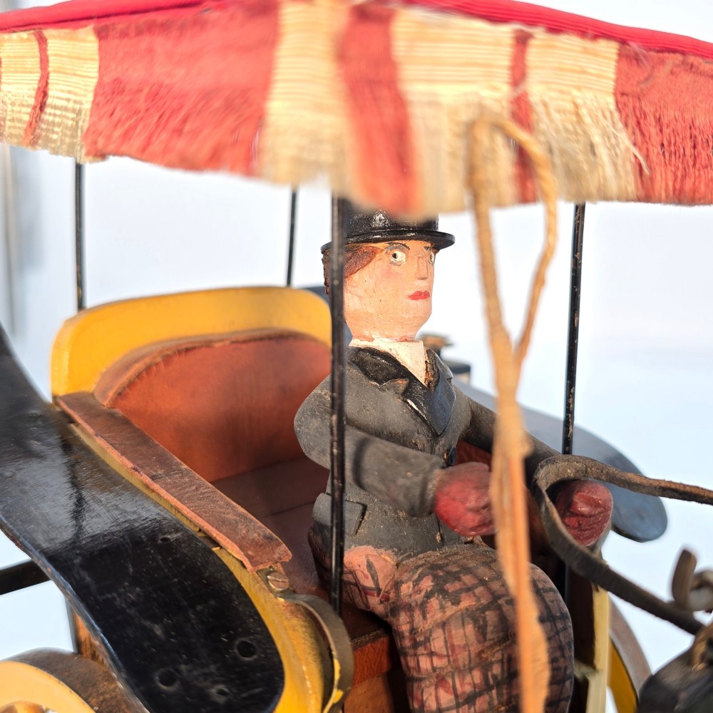 A hand-crafted 1928 wooden folk art model featuring a brown horse attached to a yellow two-wheeled chaise carriage with a driver figure under a striped canopy, mounted on a dark wooden base. View VI