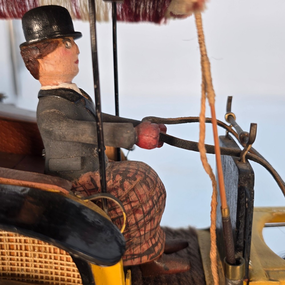 A hand-crafted 1928 wooden folk art model featuring a brown horse attached to a yellow two-wheeled chaise carriage with a driver figure under a striped canopy, mounted on a dark wooden base. View IX