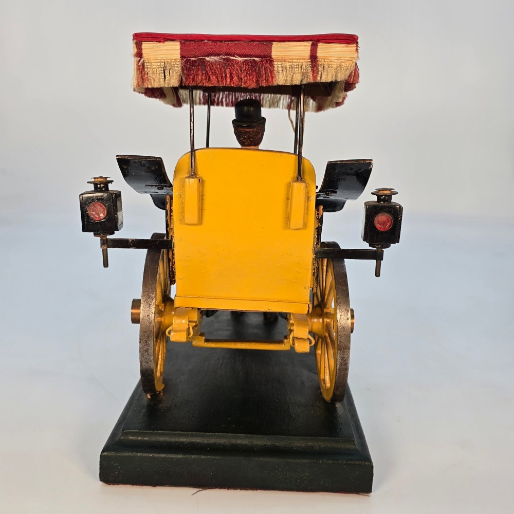 A hand-crafted 1928 wooden folk art model featuring a brown horse attached to a yellow two-wheeled chaise carriage with a driver figure under a striped canopy, mounted on a dark wooden base. View X