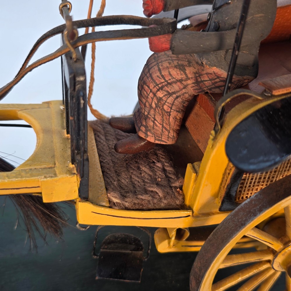A hand-crafted 1928 wooden folk art model featuring a brown horse attached to a yellow two-wheeled chaise carriage with a driver figure under a striped canopy, mounted on a dark wooden base. View XI