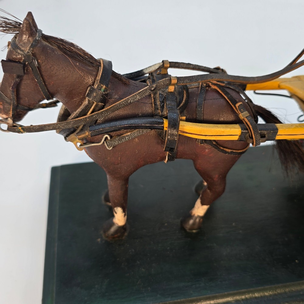 A hand-crafted 1928 wooden folk art model featuring a brown horse attached to a yellow two-wheeled chaise carriage with a driver figure under a striped canopy, mounted on a dark wooden base. View XII