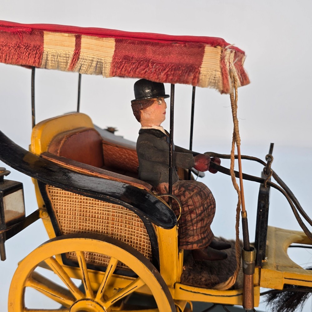 A hand-crafted 1928 wooden folk art model featuring a brown horse attached to a yellow two-wheeled chaise carriage with a driver figure under a striped canopy, mounted on a dark wooden base. View XIII