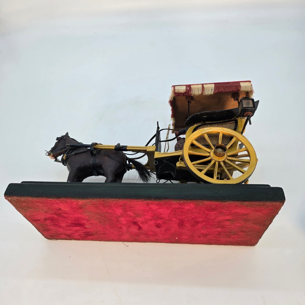 A hand-crafted 1928 wooden folk art model featuring a brown horse attached to a yellow two-wheeled chaise carriage with a driver figure under a striped canopy, mounted on a dark wooden base. View XV
