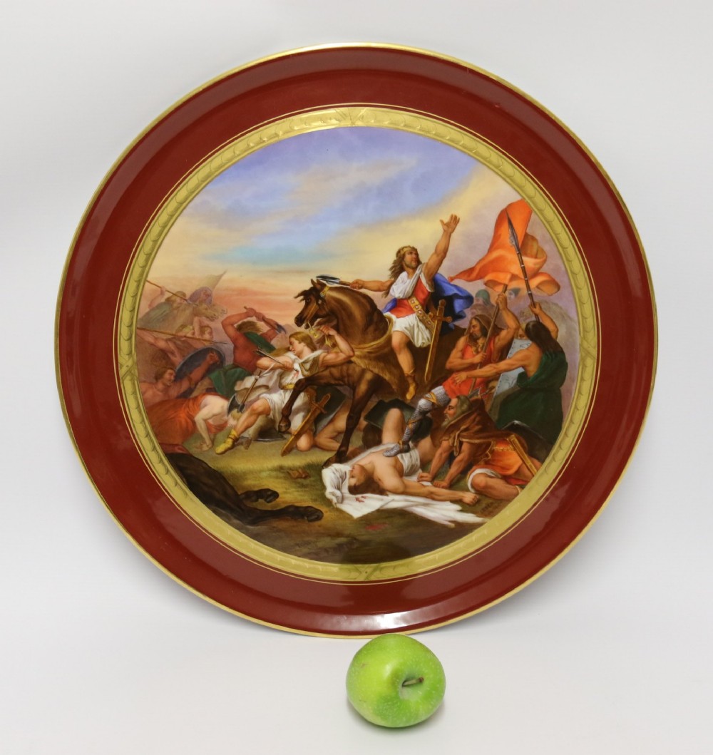 A large round porcelain charger with a deep burgundy border edged in gold leaf, featuring a central hand-painted scene depicting a historic battle with figures on horseback.