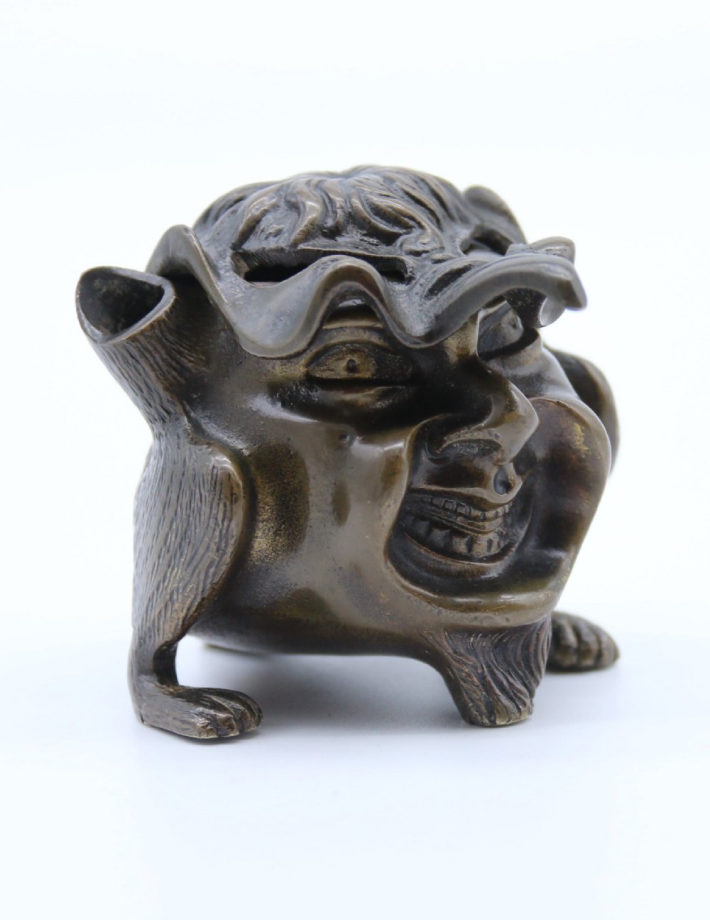 A dark cast bronze novelty inkwell sculpted in the form of a grotesque grinning face with textured hair and small footed base.