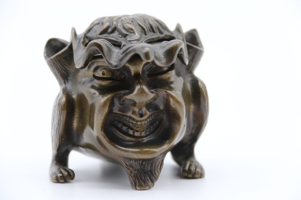 A dark cast bronze novelty inkwell sculpted in the form of a grotesque grinning face with textured hair and small footed base. View II