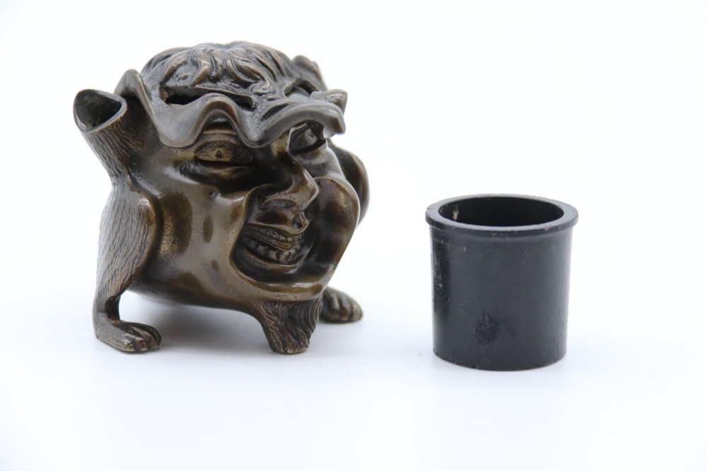 A dark cast bronze novelty inkwell sculpted in the form of a grotesque grinning face with textured hair and small footed base. View IV