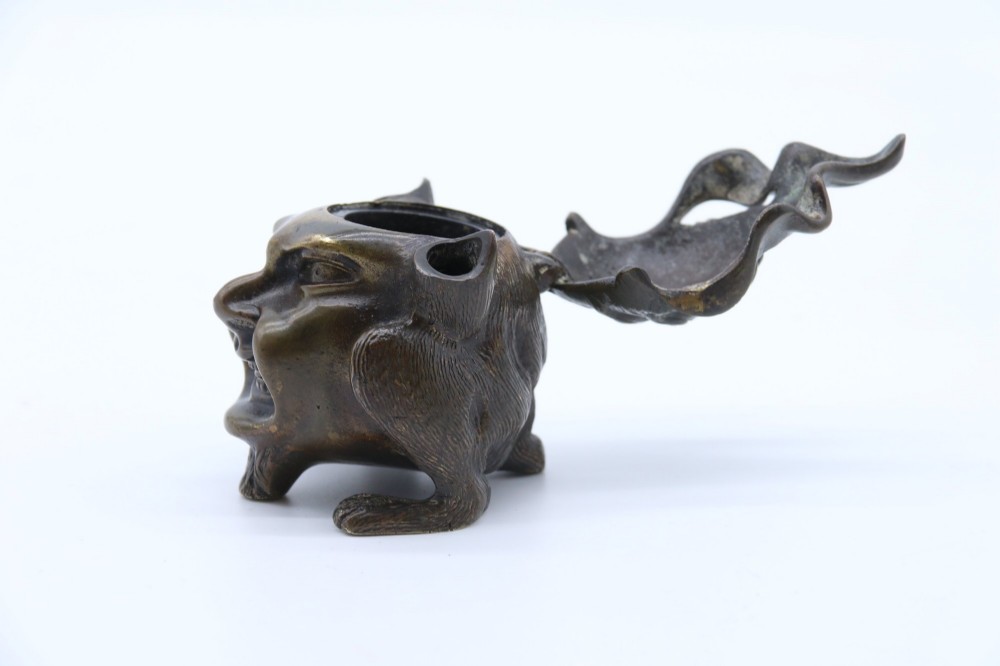 A dark cast bronze novelty inkwell sculpted in the form of a grotesque grinning face with textured hair and small footed base. View V