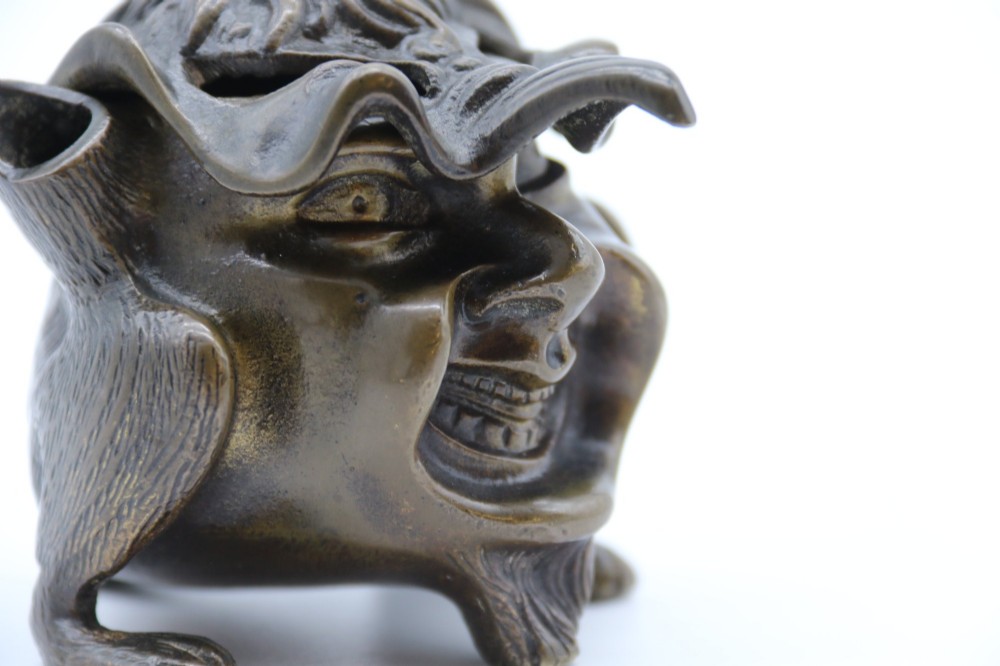 A dark cast bronze novelty inkwell sculpted in the form of a grotesque grinning face with textured hair and small footed base. View VII