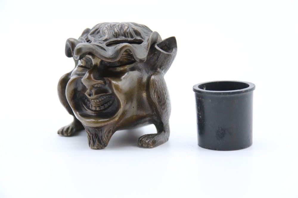 A dark cast bronze novelty inkwell sculpted in the form of a grotesque grinning face with textured hair and small footed base. View VIII