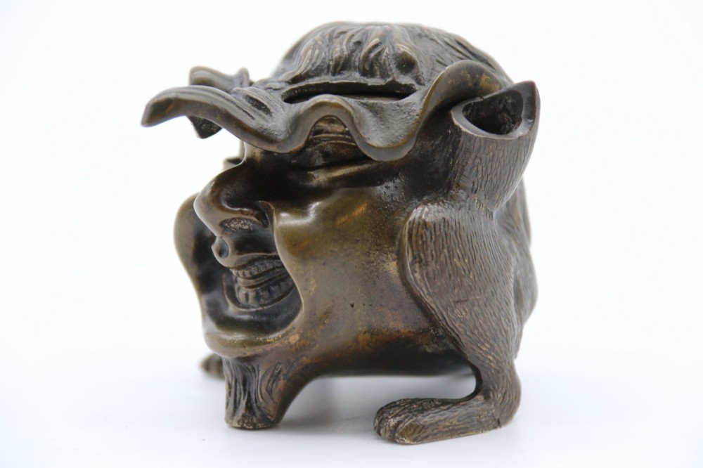 A dark cast bronze novelty inkwell sculpted in the form of a grotesque grinning face with textured hair and small footed base. View IX