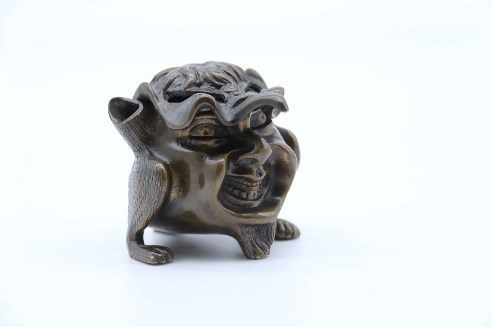 A dark cast bronze novelty inkwell sculpted in the form of a grotesque grinning face with textured hair and small footed base. View X