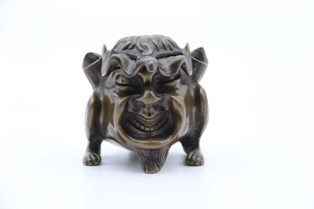 A dark cast bronze novelty inkwell sculpted in the form of a grotesque grinning face with textured hair and small footed base. View XI
