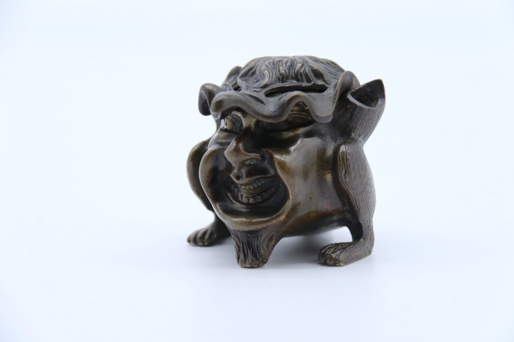 A dark cast bronze novelty inkwell sculpted in the form of a grotesque grinning face with textured hair and small footed base. View XII