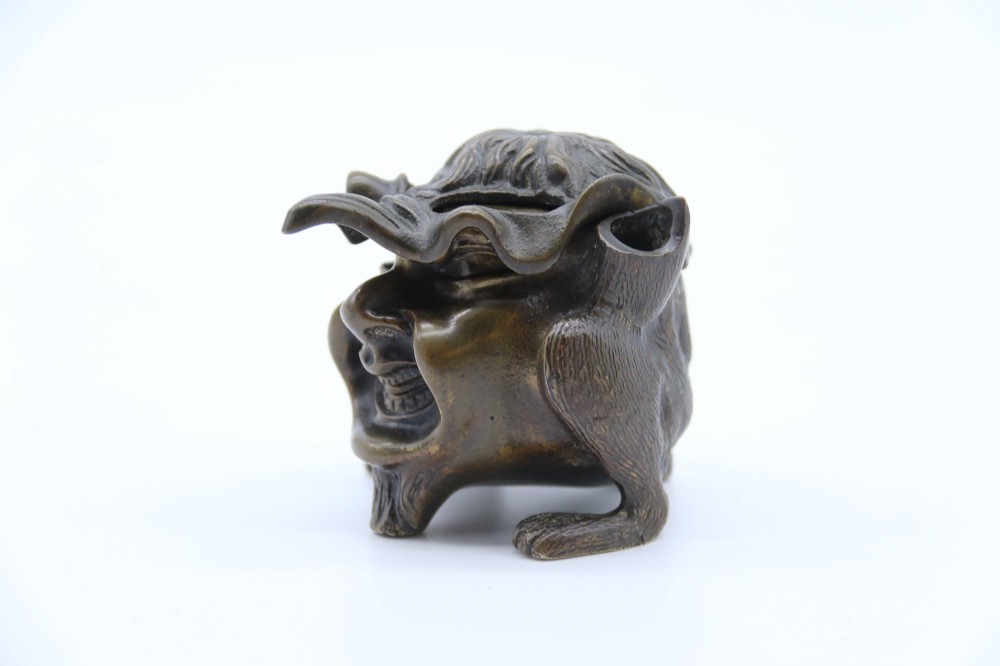 A dark cast bronze novelty inkwell sculpted in the form of a grotesque grinning face with textured hair and small footed base. View XIII