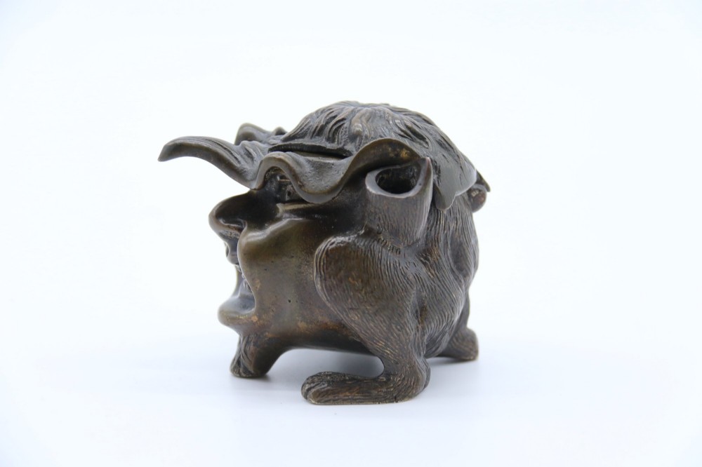 A dark cast bronze novelty inkwell sculpted in the form of a grotesque grinning face with textured hair and small footed base. View XIV