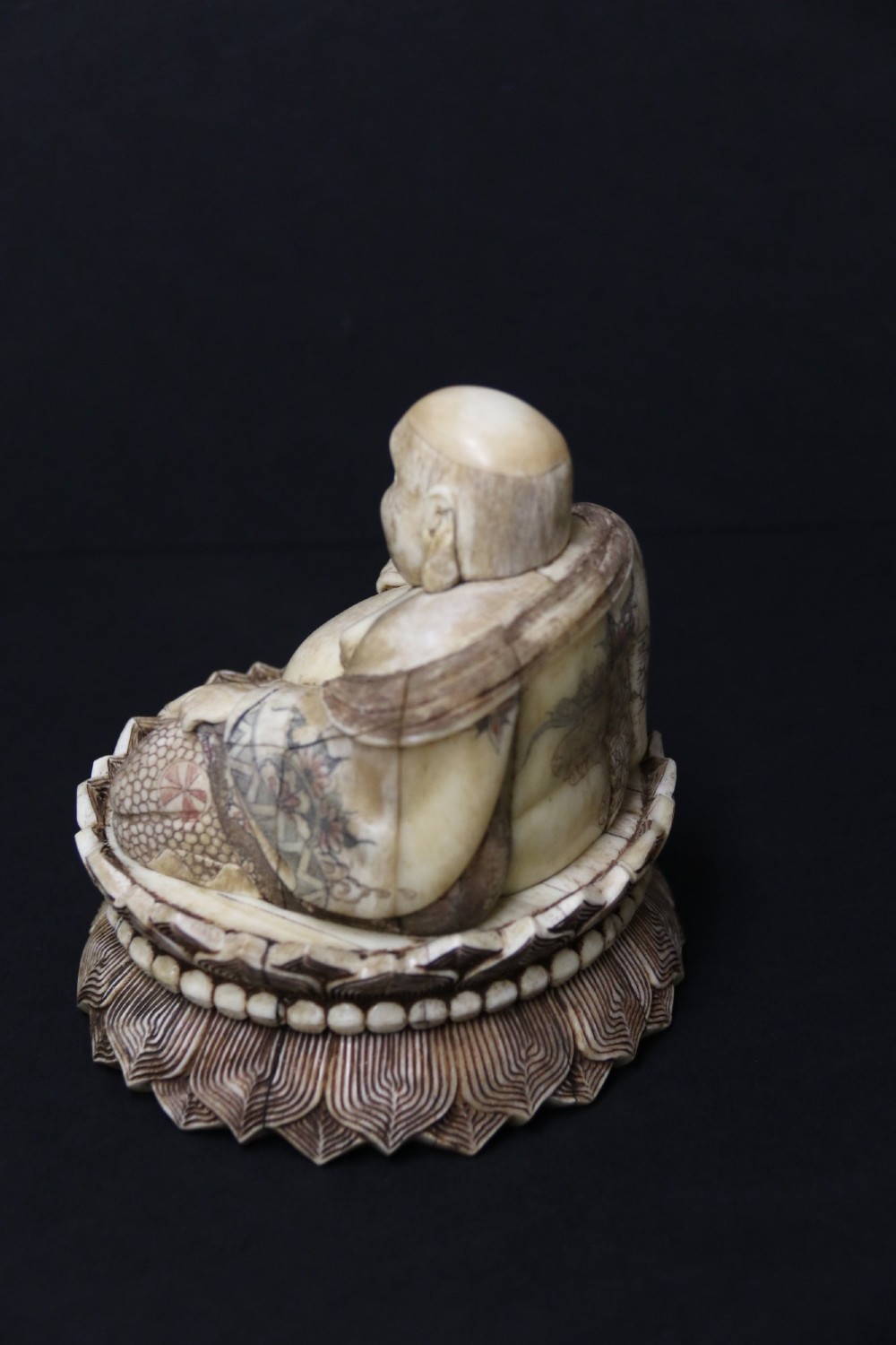 A Japanese carved bone okimono depicting a seated Buddha figure on an engraved lotus base with intricate ink detailing. View V