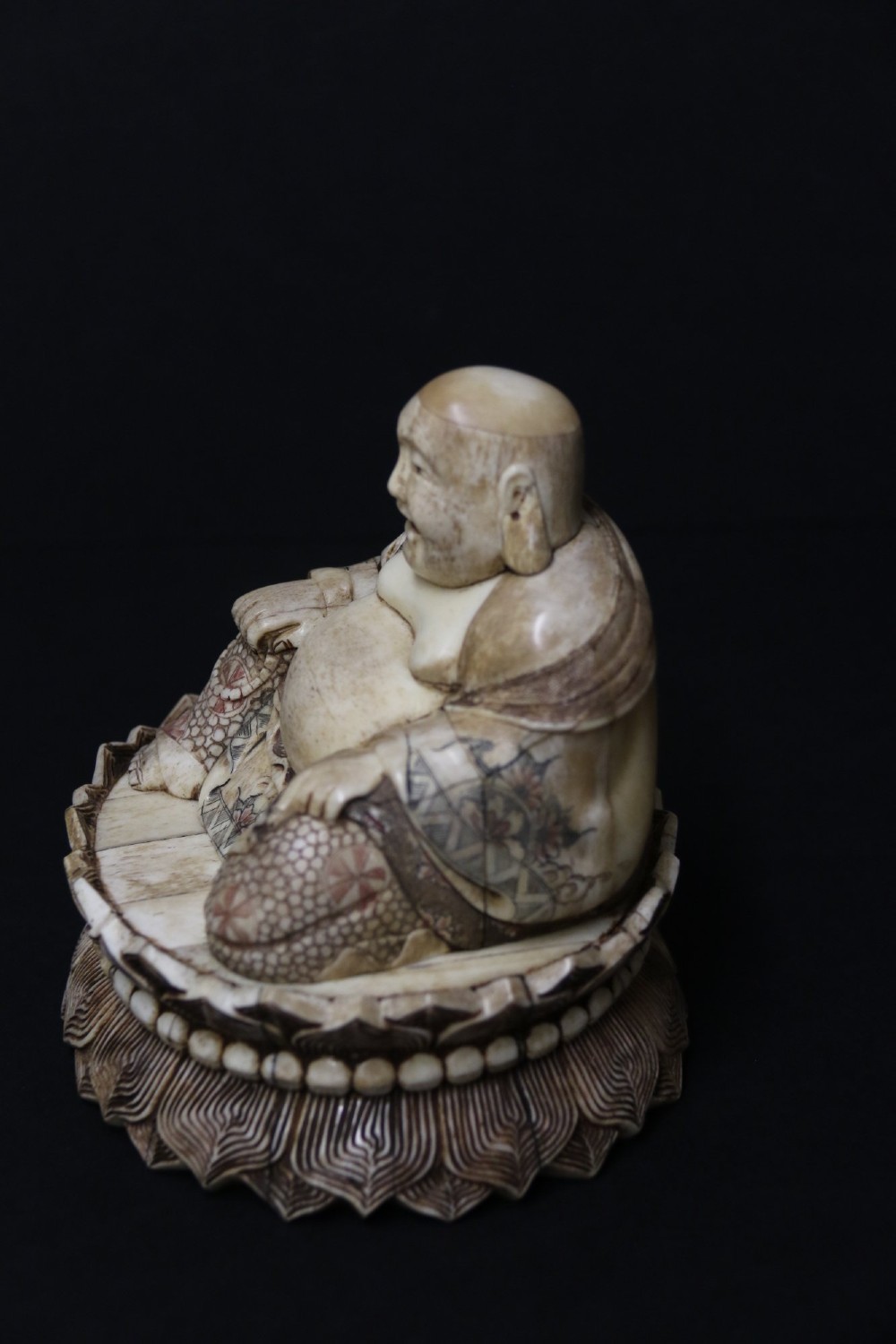 A Japanese carved bone okimono depicting a seated Buddha figure on an engraved lotus base with intricate ink detailing. View VI
