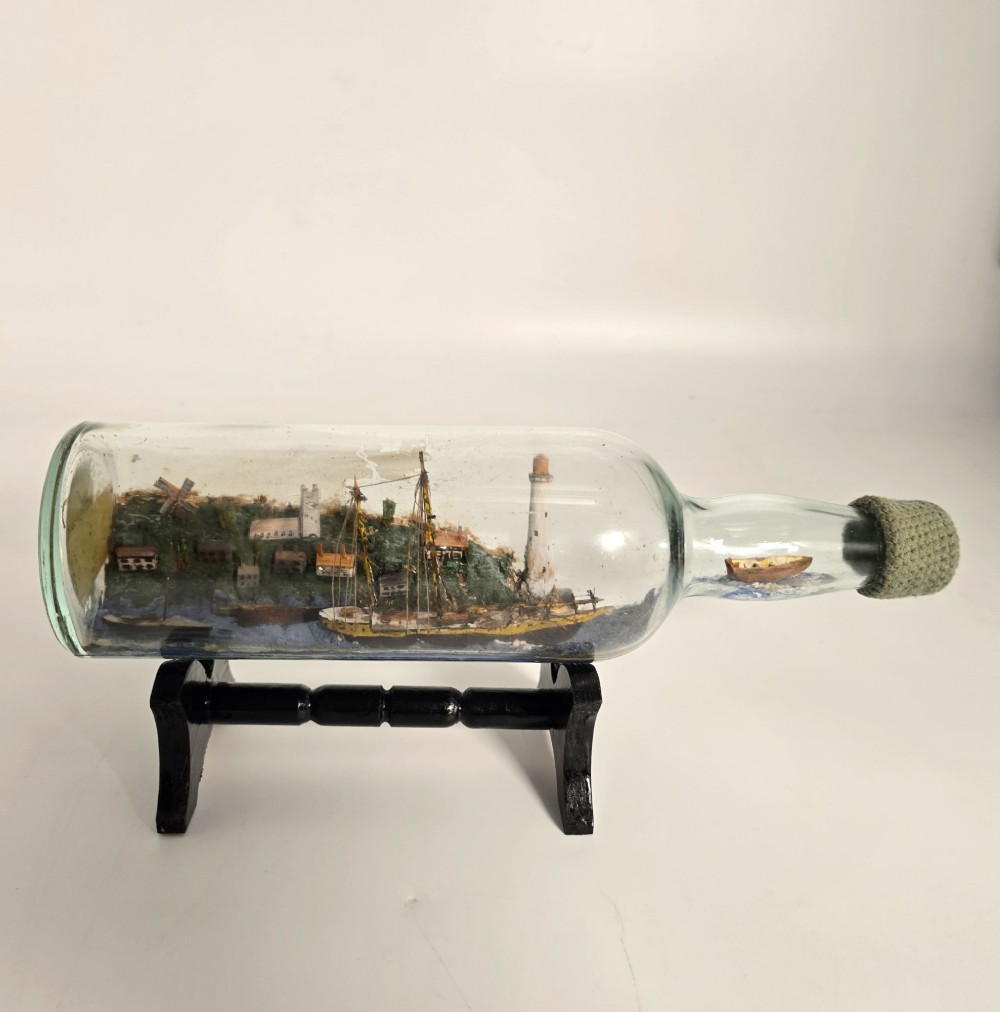 A clear glass bottle containing a handcrafted folk art diorama of a sailing vessel, lighthouse, and coastal town, supported by a black wooden display stand.