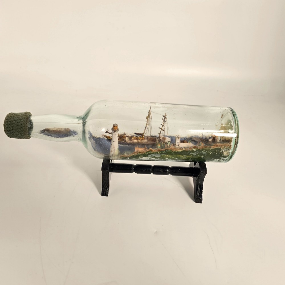 A clear glass bottle containing a handcrafted folk art diorama of a sailing vessel, lighthouse, and coastal town, supported by a black wooden display stand. View III