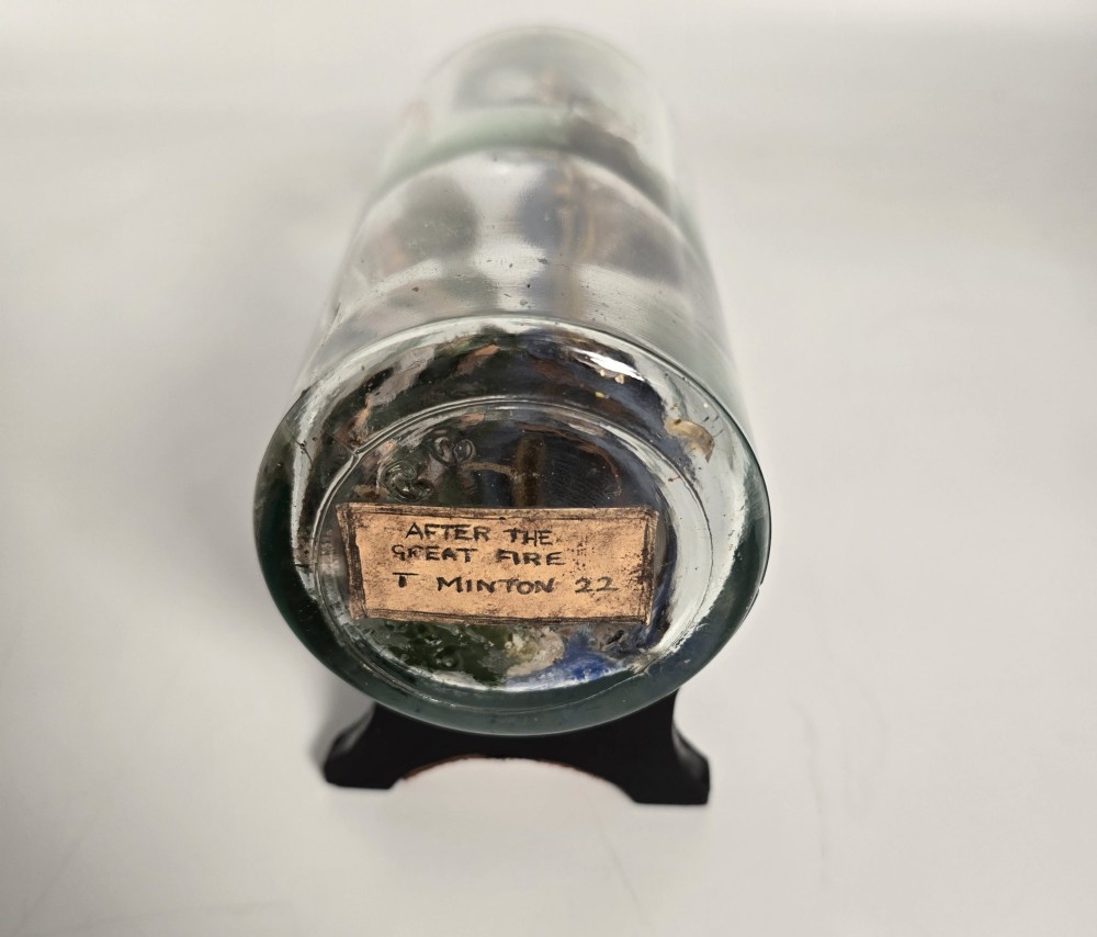 A clear glass bottle containing a handcrafted folk art diorama of a sailing vessel, lighthouse, and coastal town, supported by a black wooden display stand. View IV