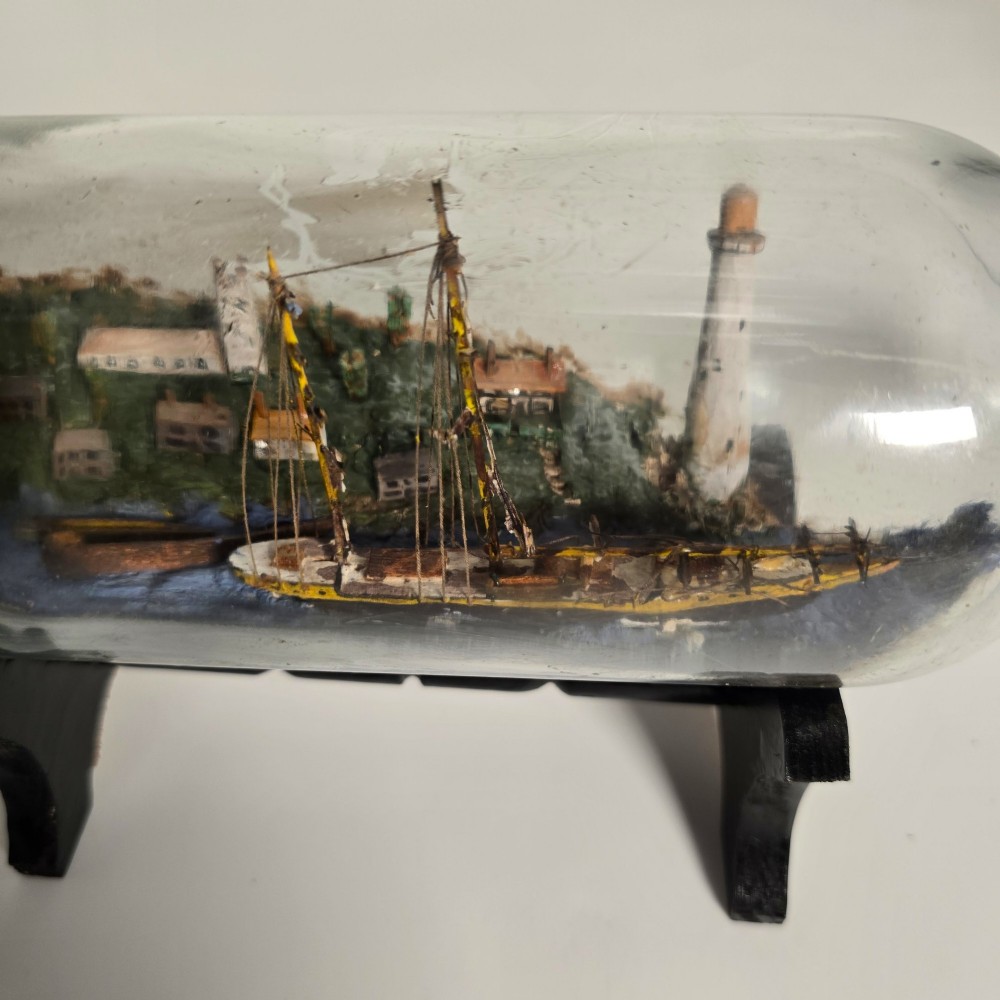 A clear glass bottle containing a handcrafted folk art diorama of a sailing vessel, lighthouse, and coastal town, supported by a black wooden display stand. View V