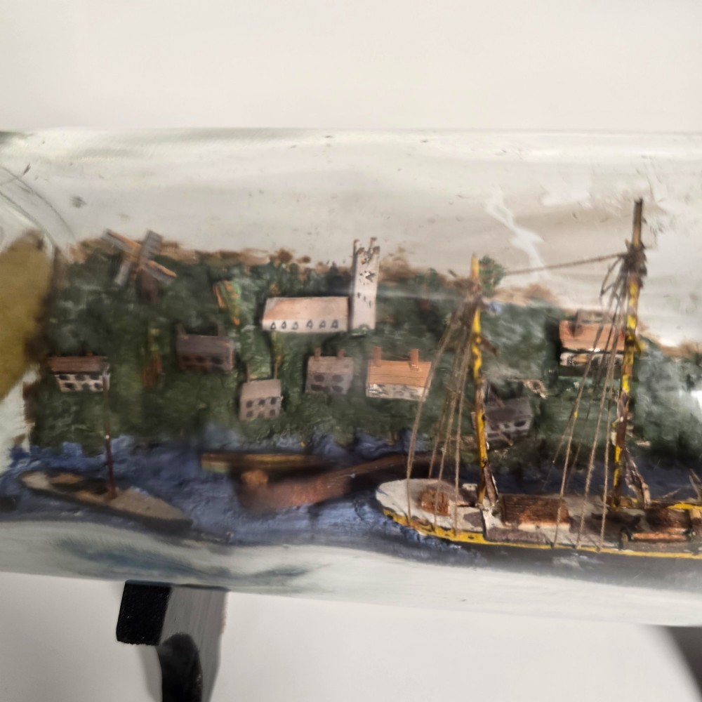 A clear glass bottle containing a handcrafted folk art diorama of a sailing vessel, lighthouse, and coastal town, supported by a black wooden display stand. View VI