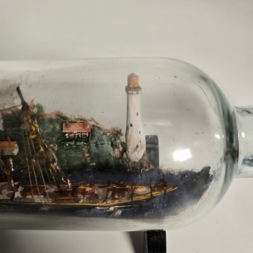 A clear glass bottle containing a handcrafted folk art diorama of a sailing vessel, lighthouse, and coastal town, supported by a black wooden display stand. View VII
