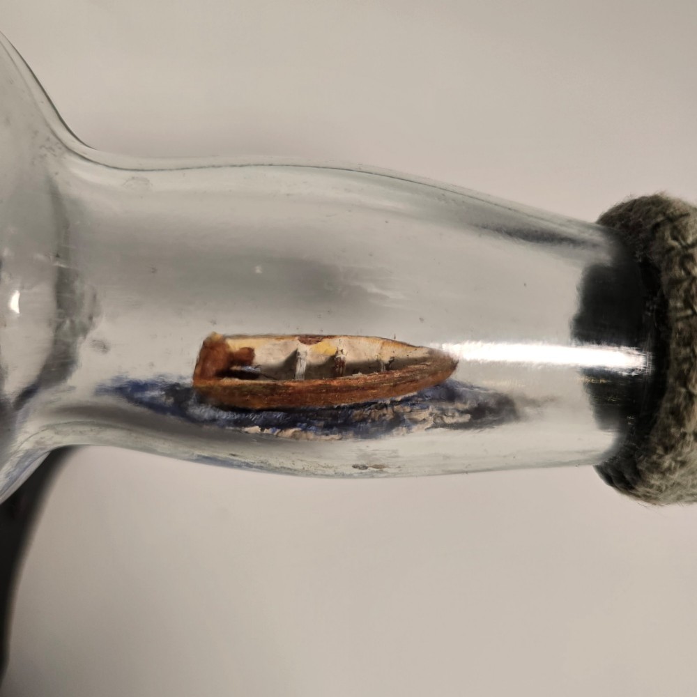 A clear glass bottle containing a handcrafted folk art diorama of a sailing vessel, lighthouse, and coastal town, supported by a black wooden display stand. View VIII