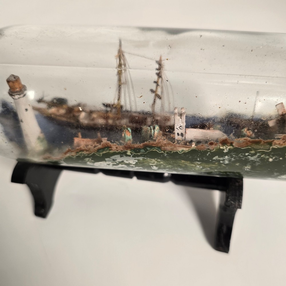 A clear glass bottle containing a handcrafted folk art diorama of a sailing vessel, lighthouse, and coastal town, supported by a black wooden display stand. View X