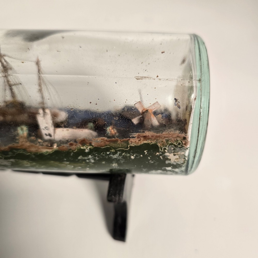 A clear glass bottle containing a handcrafted folk art diorama of a sailing vessel, lighthouse, and coastal town, supported by a black wooden display stand. View XI