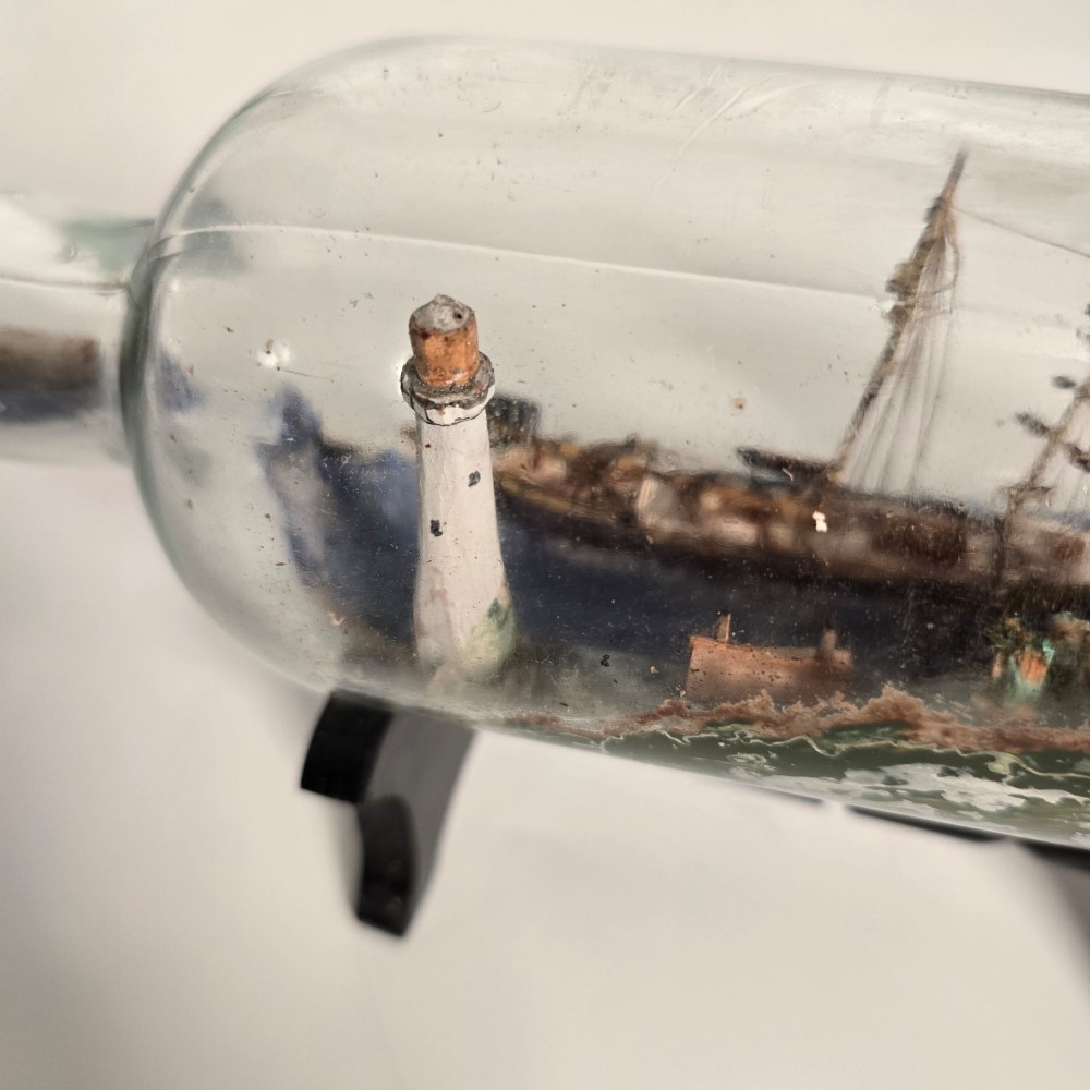 A clear glass bottle containing a handcrafted folk art diorama of a sailing vessel, lighthouse, and coastal town, supported by a black wooden display stand. View XII