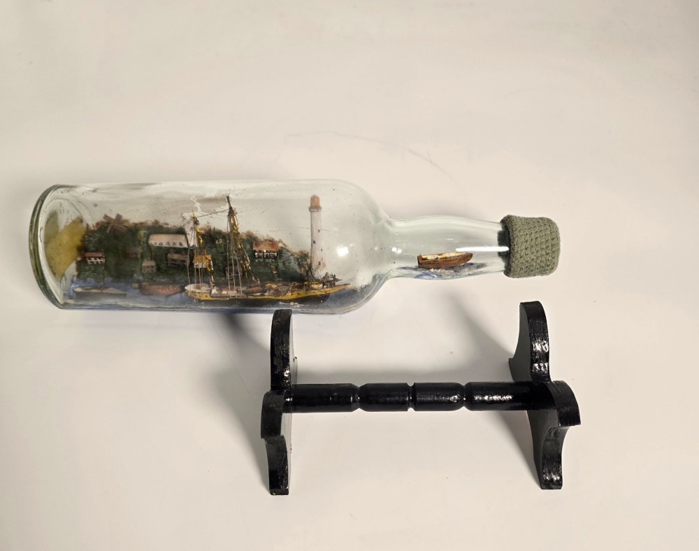 A clear glass bottle containing a handcrafted folk art diorama of a sailing vessel, lighthouse, and coastal town, supported by a black wooden display stand. View XIII