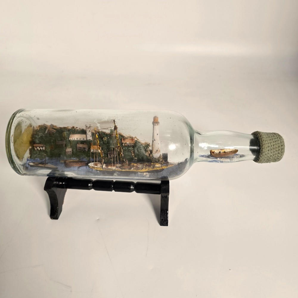 A clear glass bottle containing a handcrafted folk art diorama of a sailing vessel, lighthouse, and coastal town, supported by a black wooden display stand. View XIV