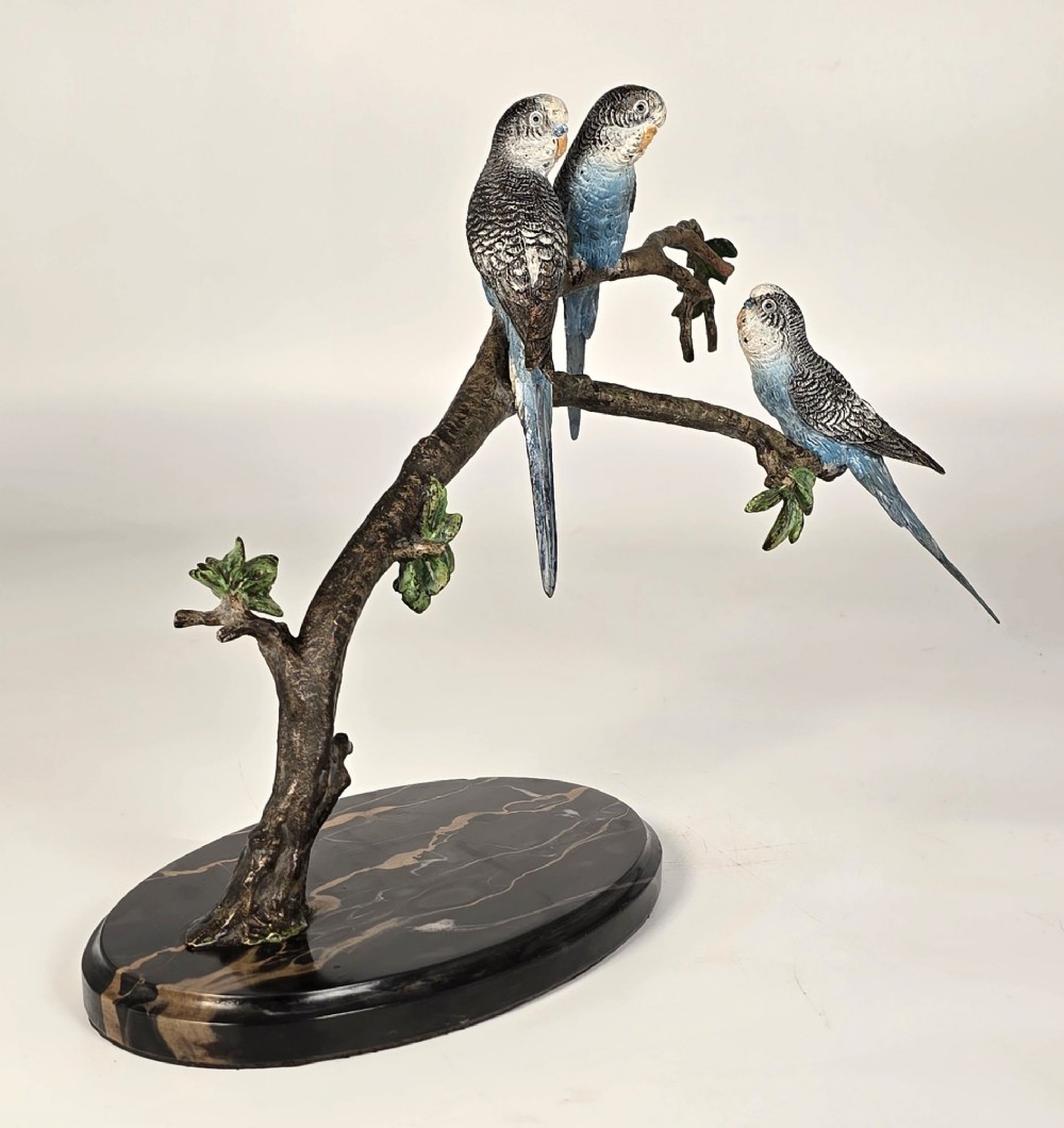 A cold-painted bronze sculpture featuring three blue and grey budgerigars perched on a leafy bronze branch, mounted on an oval dark marble base.