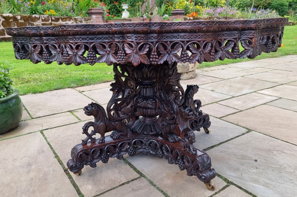 A mid-19th century Anglo-Indian padauk centre table featuring a scalloped edge top, deeply carved pierced floral apron, and a central pedestal supported by carved lion figures on castors. View II