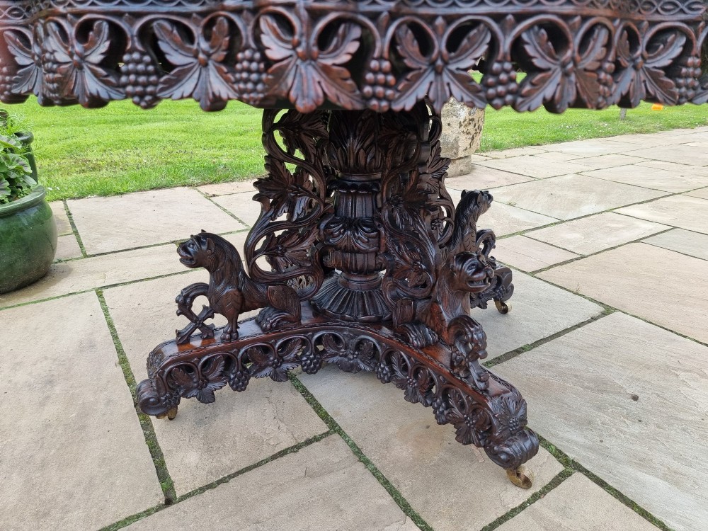 A mid-19th century Anglo-Indian padauk centre table featuring a scalloped edge top, deeply carved pierced floral apron, and a central pedestal supported by carved lion figures on castors. View V