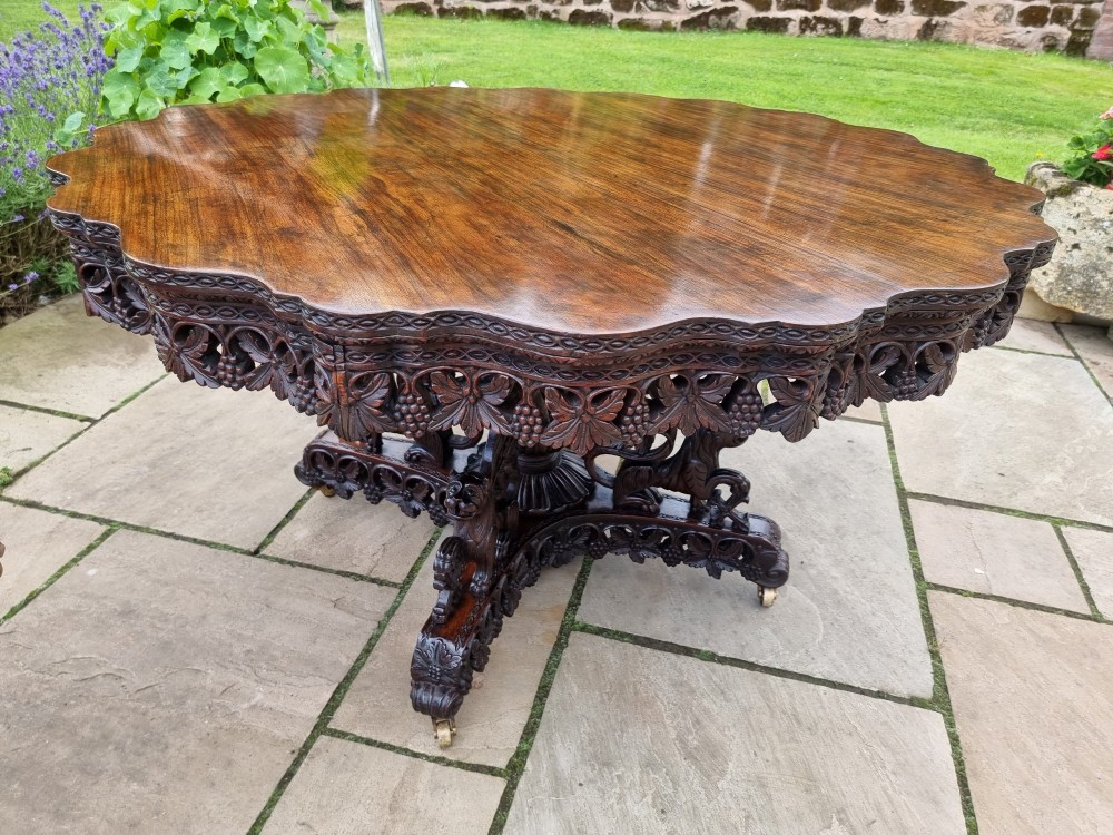 A mid-19th century Anglo-Indian padauk centre table featuring a scalloped edge top, deeply carved pierced floral apron, and a central pedestal supported by carved lion figures on castors. View VII