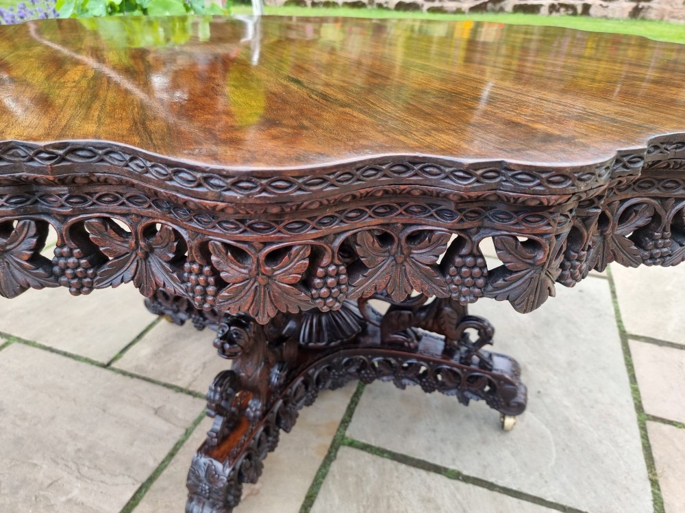 A mid-19th century Anglo-Indian padauk centre table featuring a scalloped edge top, deeply carved pierced floral apron, and a central pedestal supported by carved lion figures on castors. View VIII