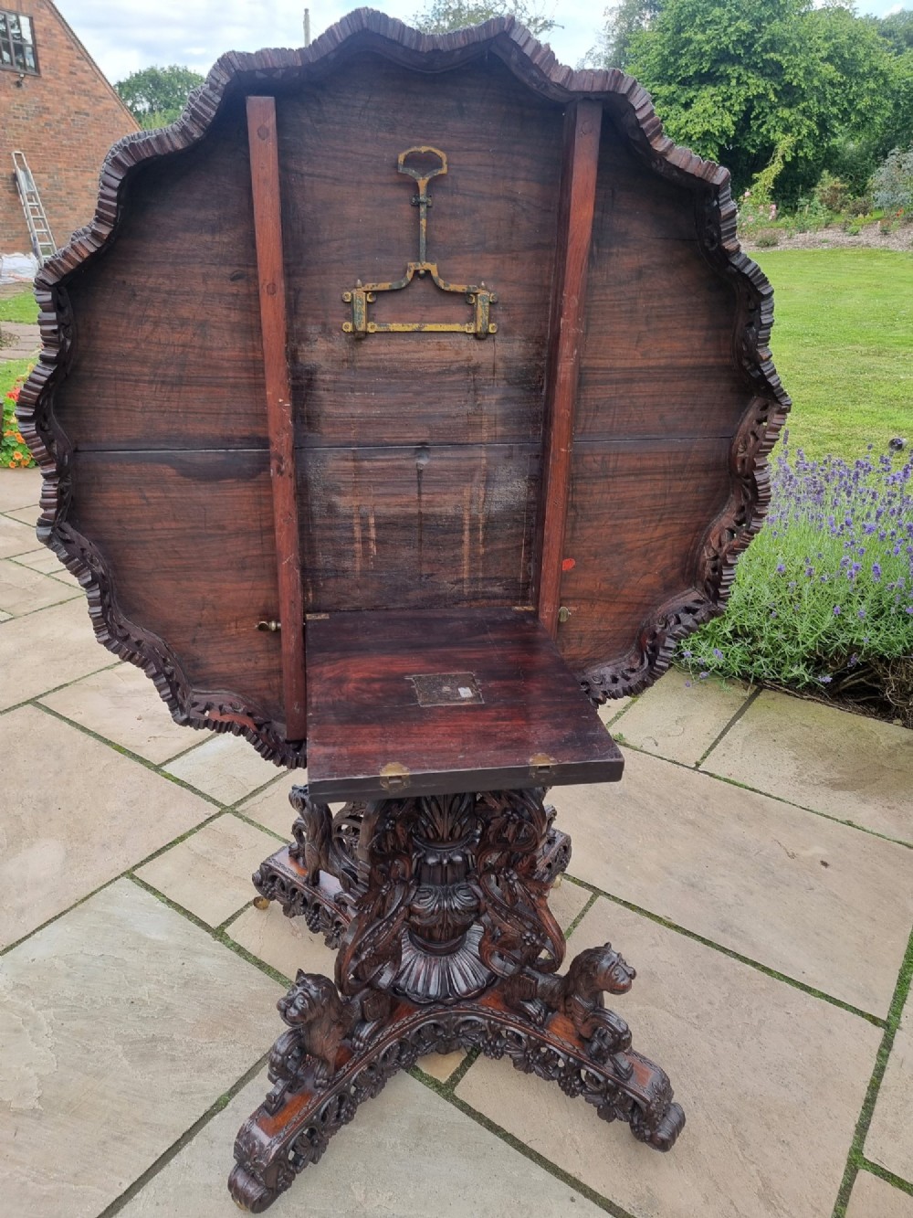 A mid-19th century Anglo-Indian padauk centre table featuring a scalloped edge top, deeply carved pierced floral apron, and a central pedestal supported by carved lion figures on castors. View IX