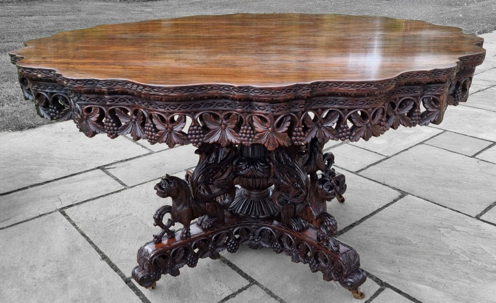A mid-19th century Anglo-Indian padauk centre table featuring a scalloped edge top, deeply carved pierced floral apron, and a central pedestal supported by carved lion figures on castors. View XV