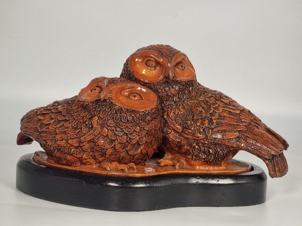 A hand-sculpted warm brown pottery pair of owls featuring textured feather detailing resting on an oval ebonised wooden base.