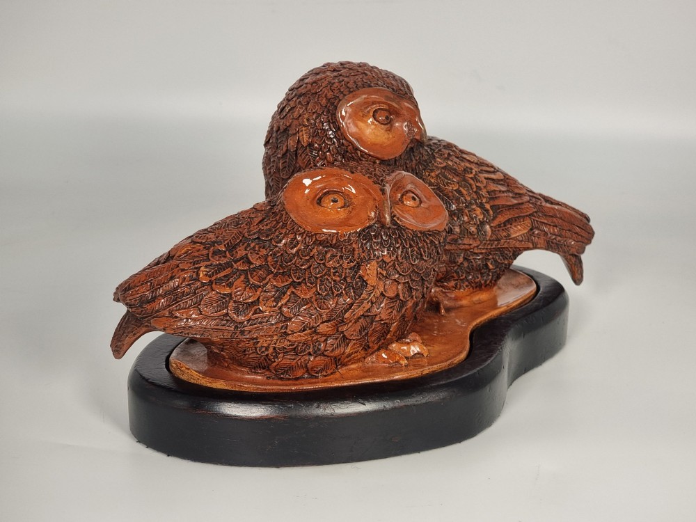 A hand-sculpted warm brown pottery pair of owls featuring textured feather detailing resting on an oval ebonised wooden base. View II
