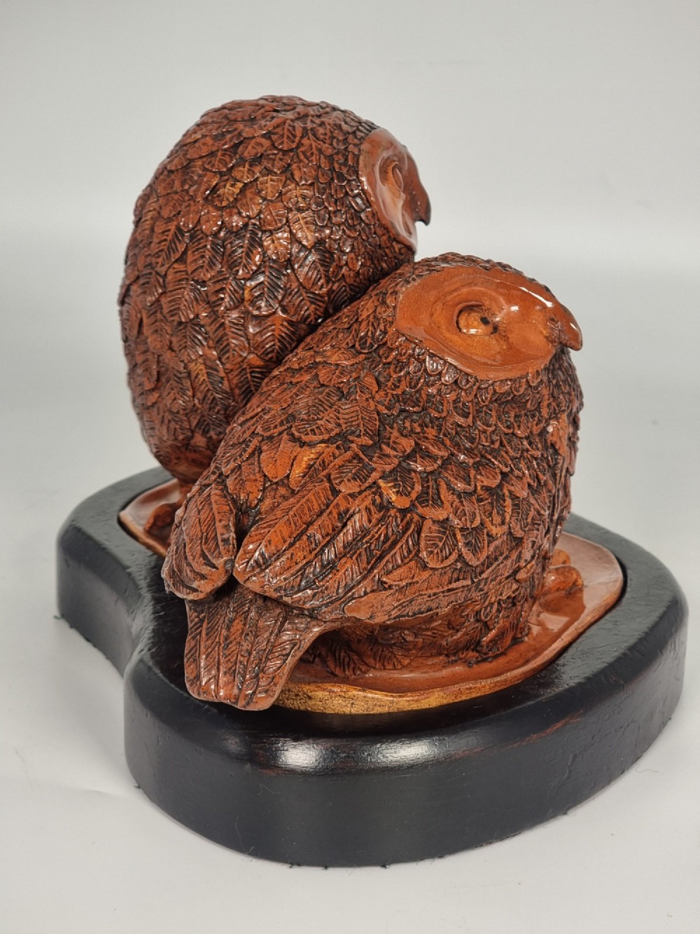 A hand-sculpted warm brown pottery pair of owls featuring textured feather detailing resting on an oval ebonised wooden base. View III