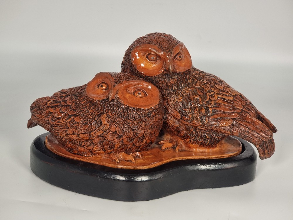 A hand-sculpted warm brown pottery pair of owls featuring textured feather detailing resting on an oval ebonised wooden base. View XV