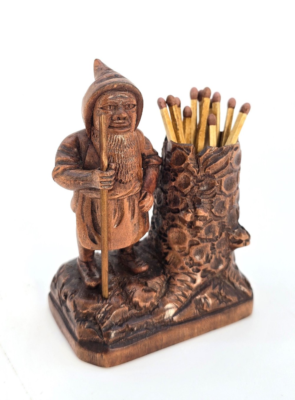 A carved brown limewood match holder depicting a hooded bearded gnome standing beside a textured tree stump container.