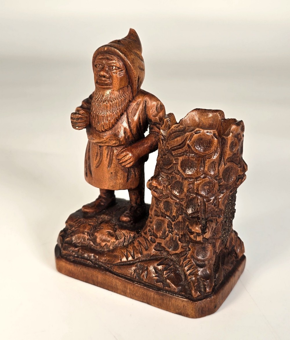 A carved brown limewood match holder depicting a hooded bearded gnome standing beside a textured tree stump container. View II