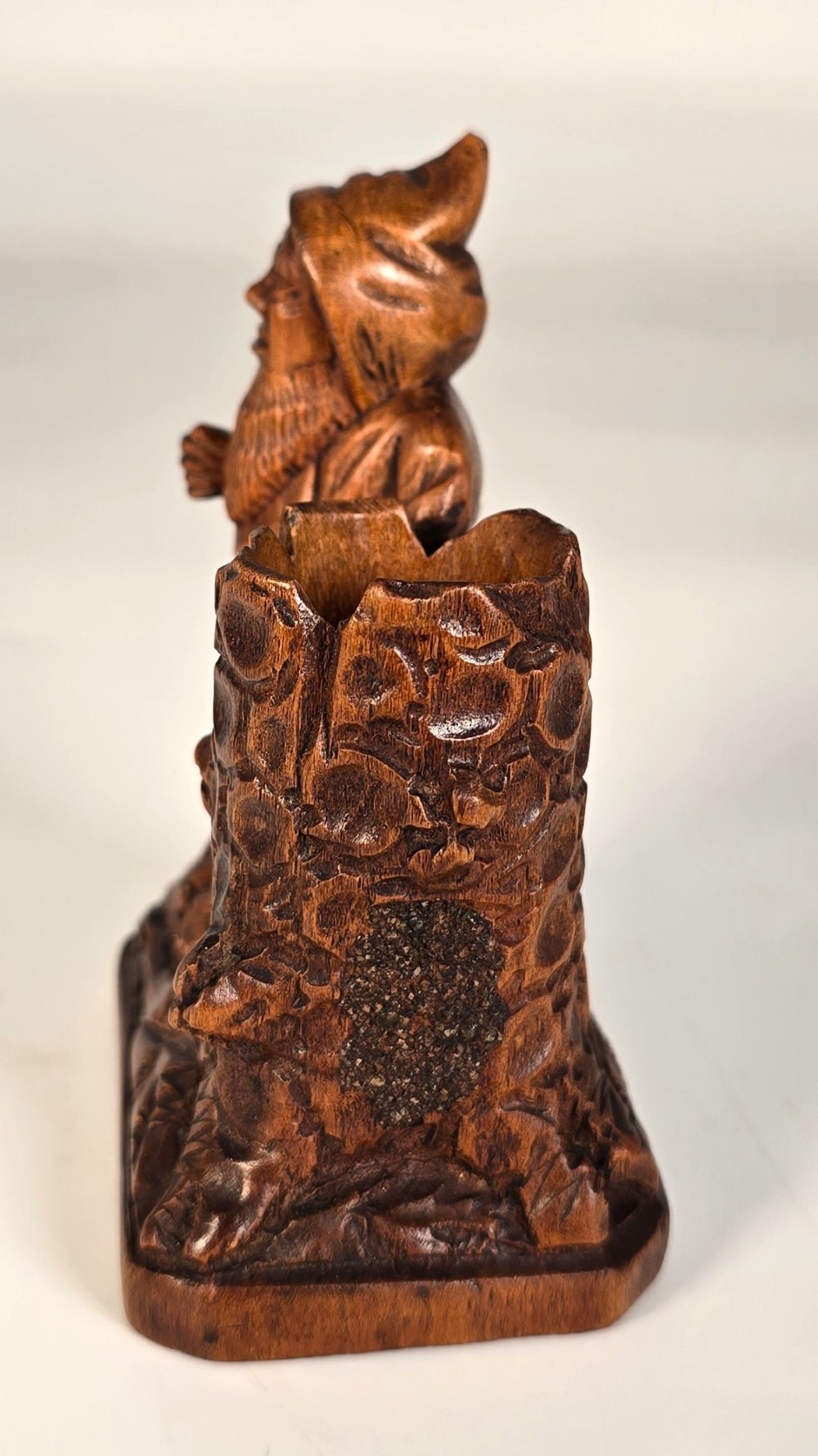 A carved brown limewood match holder depicting a hooded bearded gnome standing beside a textured tree stump container. View III